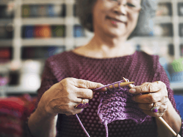 Senior woman smiling while knitting with purple yarn in a cozy environment.