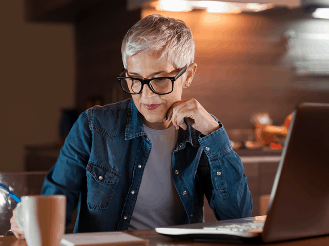 Senior woman with short hair and glasses working on a laptop in a cozy kitchen.