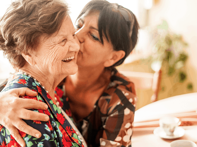 Caregiver hugging and kissing an elderly woman smiling and happy at a table.