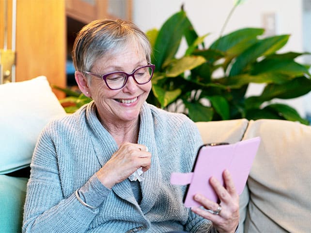Smiling senior woman with glasses using a tablet for a video call while sitting on a couch.