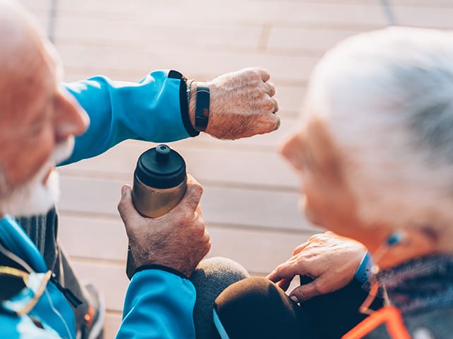 Two senior individuals viewing a smartwatch while resting outdoors with a water bottle.