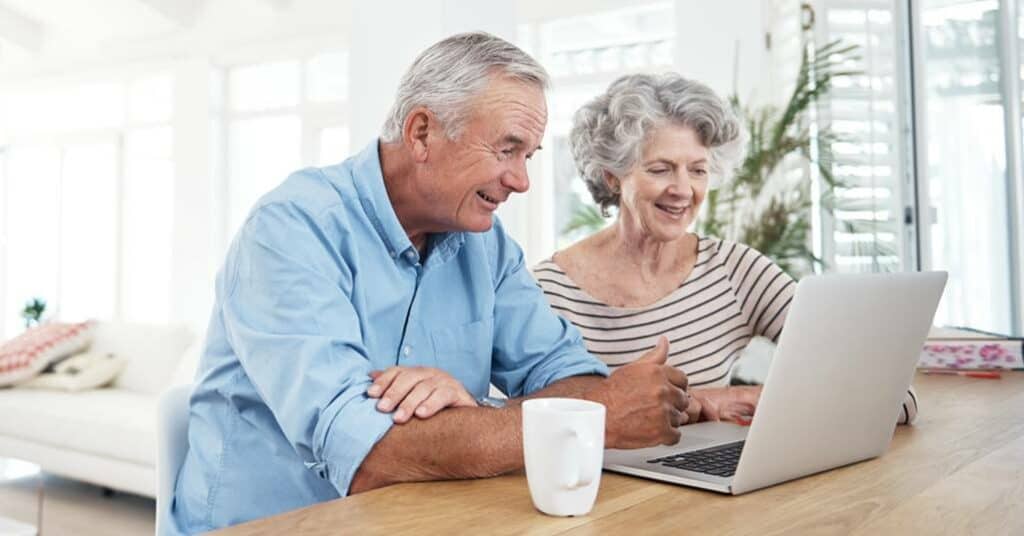 Smiling senior couple using a laptop together at a wooden table with a mug.