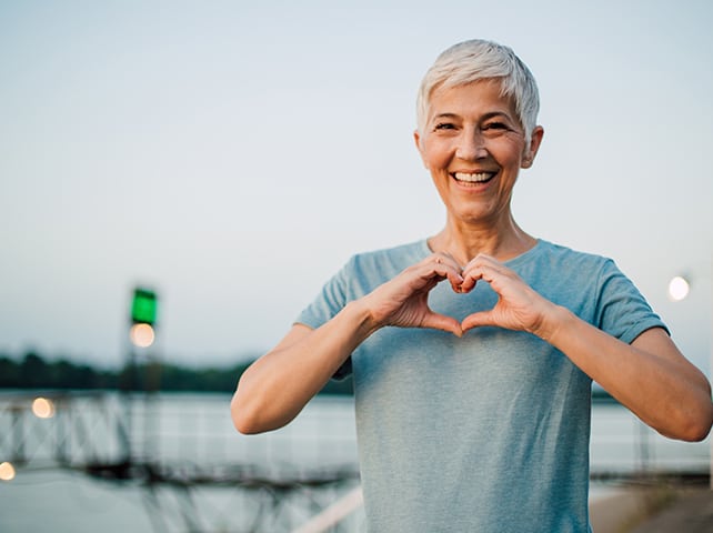 Smiling senior woman making heart shape with hands, standing outdoors by a lake.