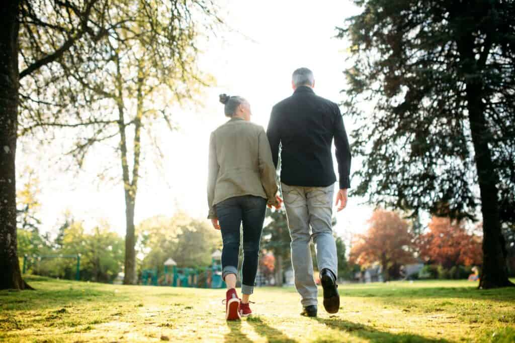 Two senior individuals holding hands and walking in a sunlit park surrounded by trees.