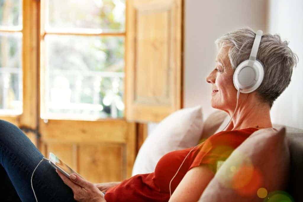 Elderly woman relaxing on couch with headphones, enjoying music in a bright room.