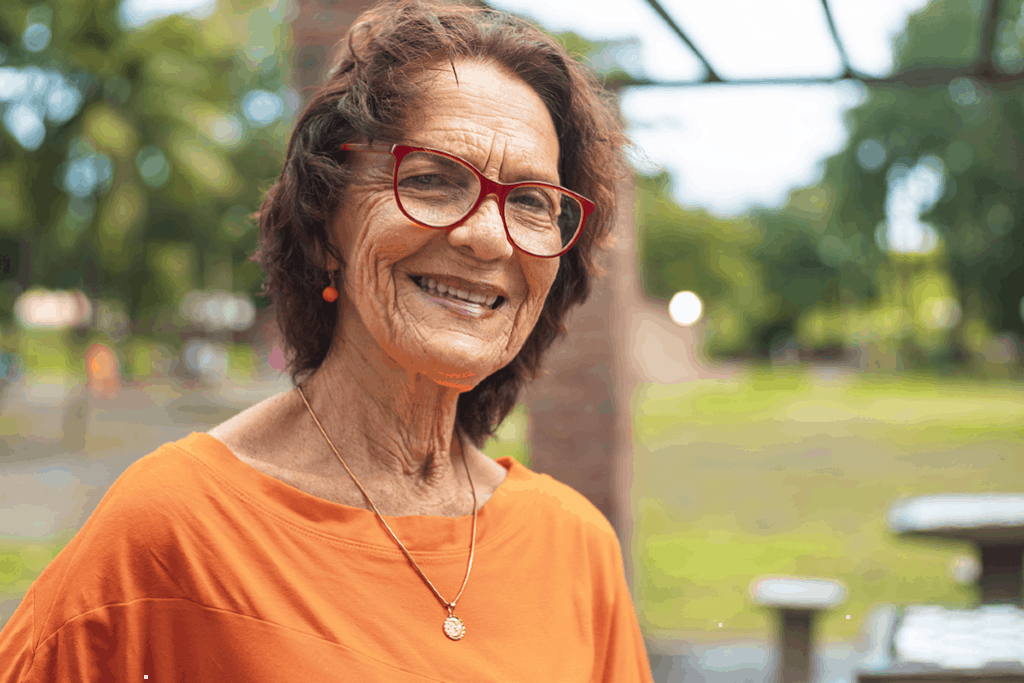 Smiling elderly woman with red glasses and orange top standing outdoors in a green area.