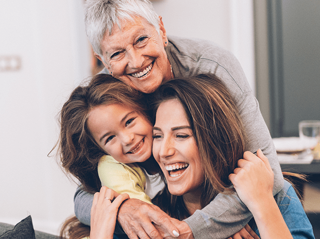 Three generations of women, grandmother, daughter, and granddaughter, smiling together indoors.
