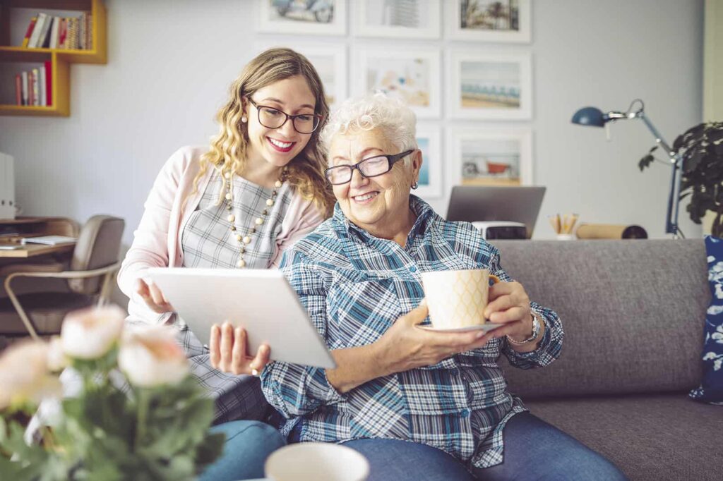 Young woman helps elderly woman use a tablet while sitting on a couch, both smiling.