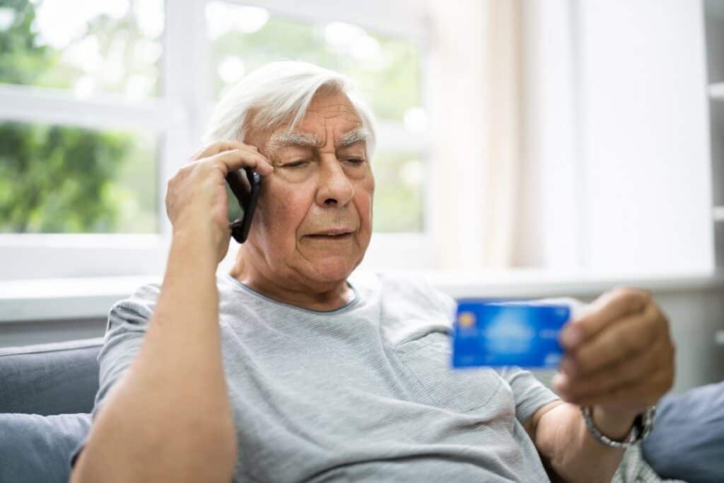 Elderly man holding a credit card and talking on the phone in a well-lit living room.