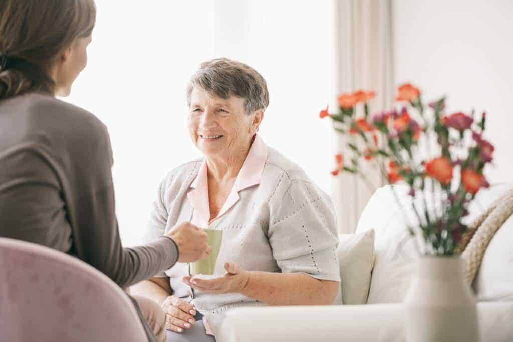 Elderly woman smiling while talking to a caretaker in a living space with flowers.