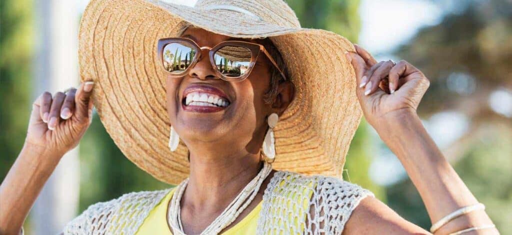 Smiling elderly woman wearing a sun hat and sunglasses outdoors on a sunny day.