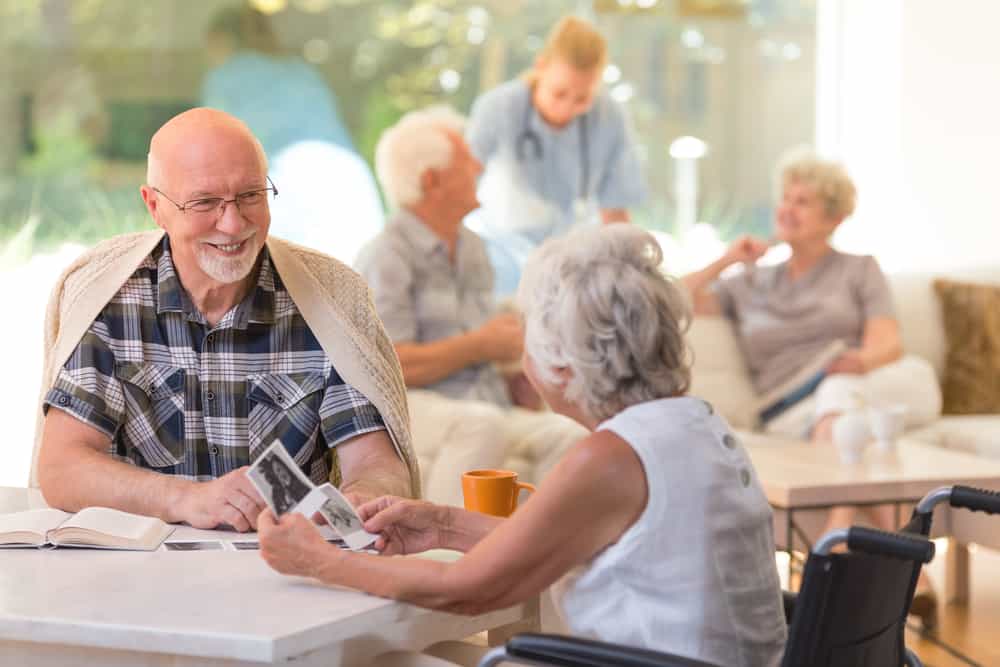 Older adults socializing and smiling in a community lounge area, with a caregiver in the background.