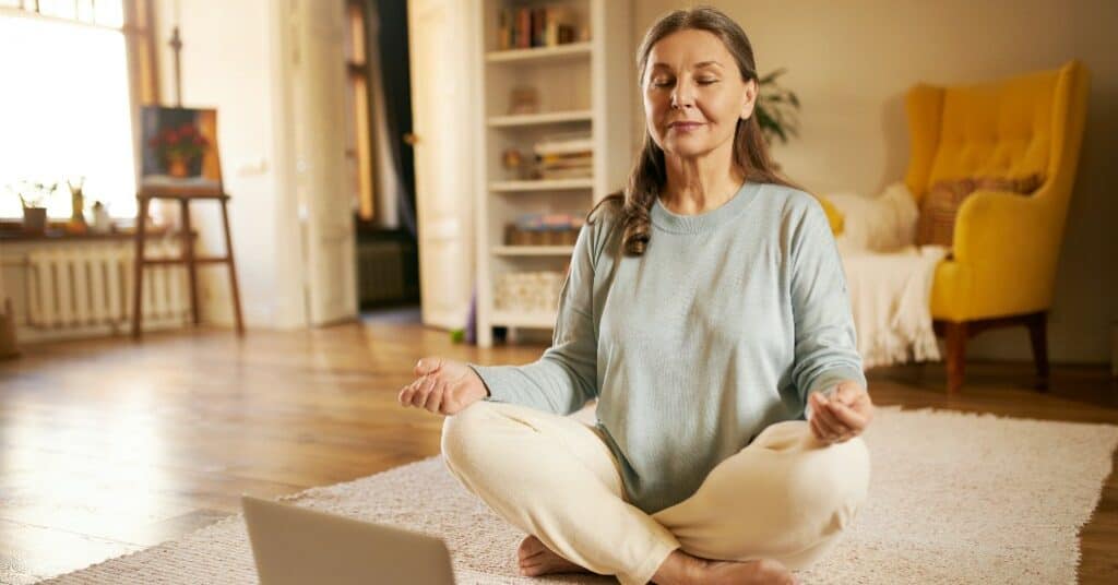 Senior woman meditating in a cozy living room with natural light and comfortable furnishings.