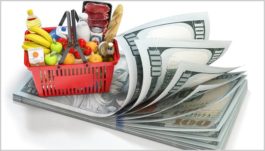 A red grocery basket filled with food items placed on a stack of hundred-dollar bills.