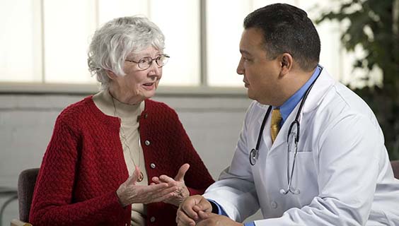 Elderly woman in red cardigan talking to a doctor in white coat with stethoscope.