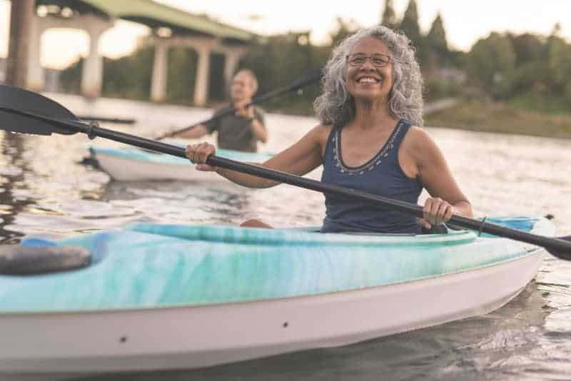 Older woman kayaking on a calm river with a smile and another person in a kayak in the background.