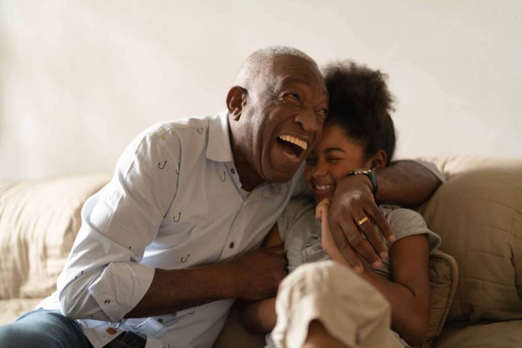 Elderly man laughing and hugging a young girl on a couch in a senior living unit.
