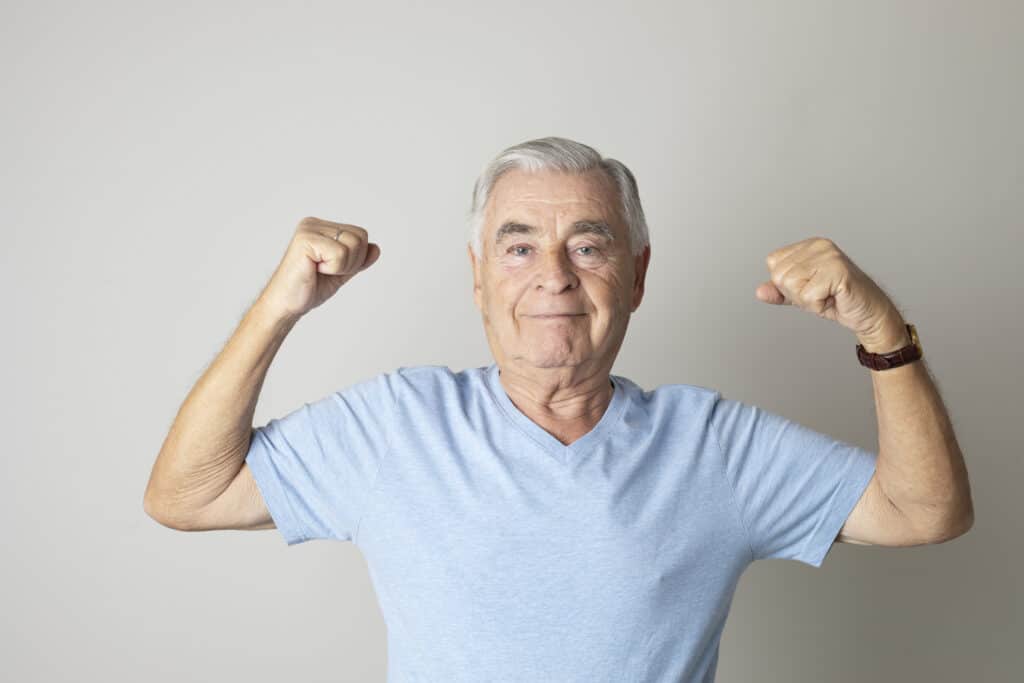 Senior man is showing his muscles to camera with raised arms.