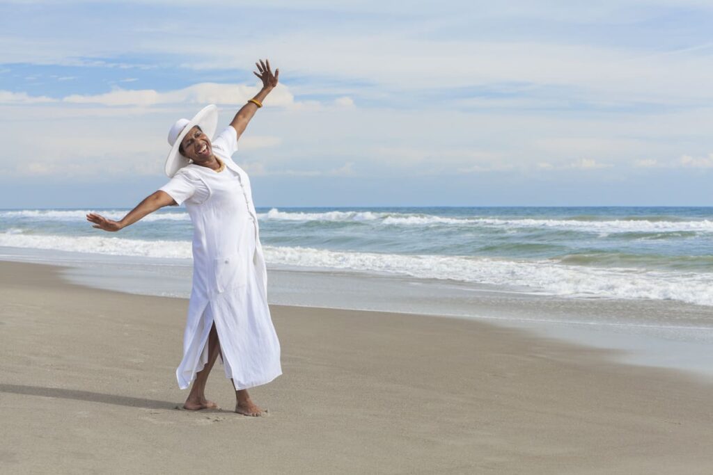 Elderly woman in white dress and hat joyfully posing on a sunny beach with ocean waves.