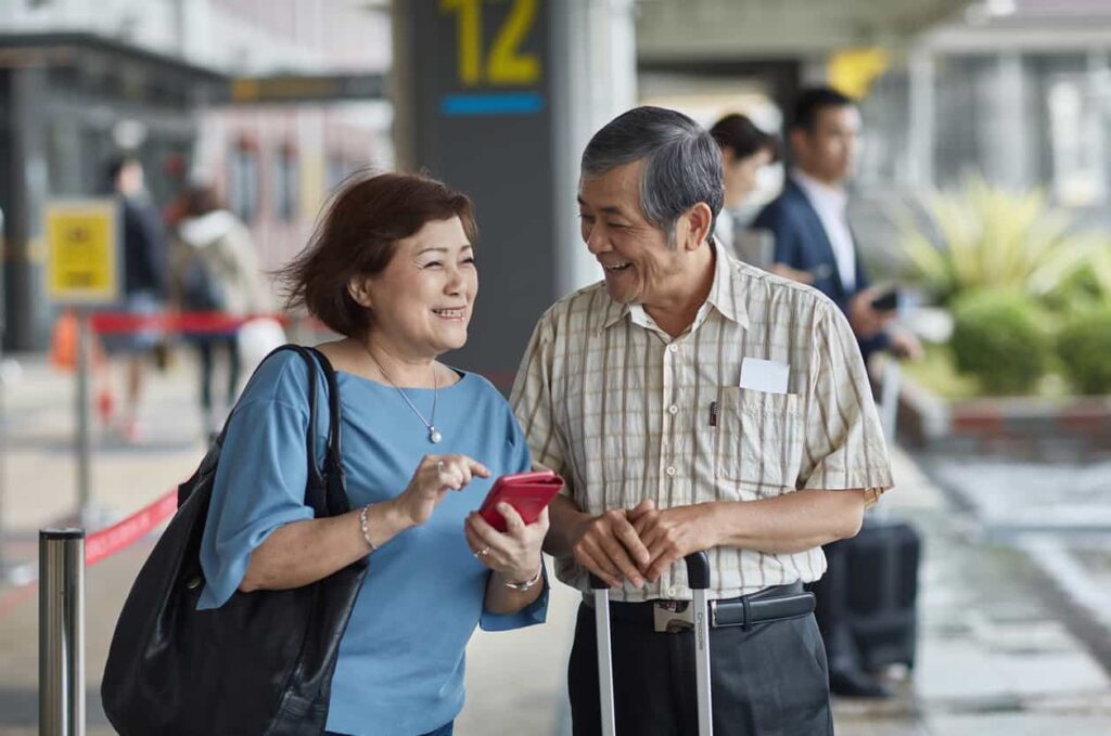 Smiling senior couple chatting near an entrance, holding a red phone and a suitcase.