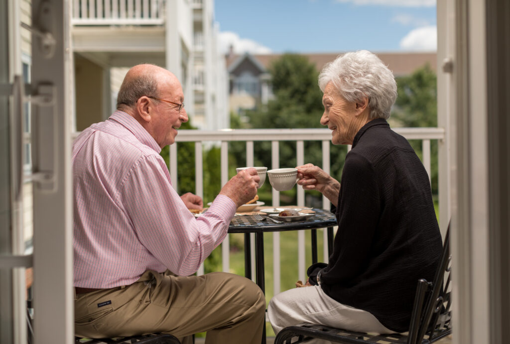 Two seniors enjoying tea on a balcony at a retirement community on a sunny day.