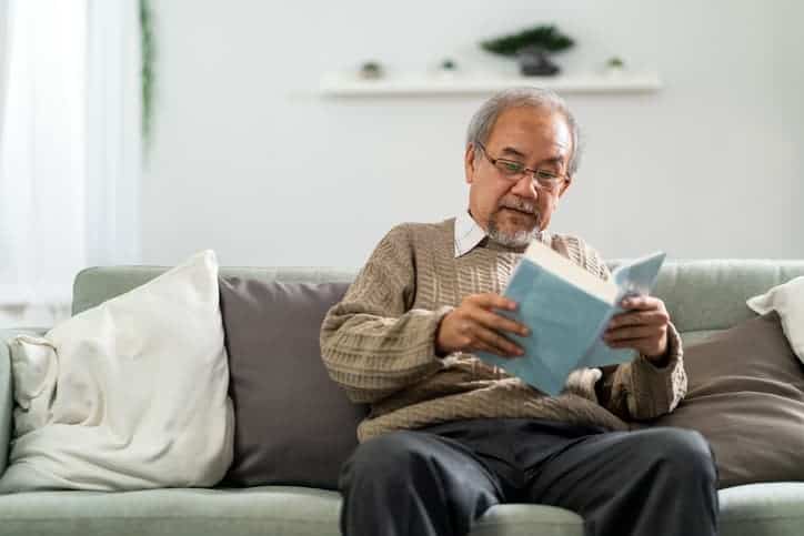 Senior man reading a book while sitting on a sofa in a living space with light furnishings.