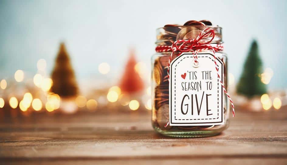 Glass jar filled with coins and a label reading Tis the season to give, surrounded by festive lights.