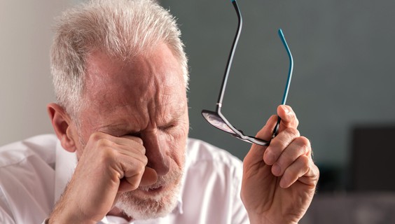 Older adult rubbing eyes while holding glasses, appearing fatigued.