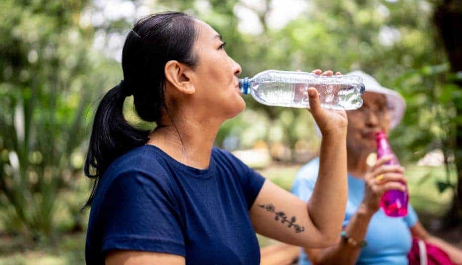 Woman drinks water from a bottle while another person does the same in a sunny outdoor setting.