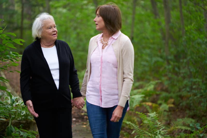 Senior woman holding hands with a loved one, walking on a wooded path and conversing.