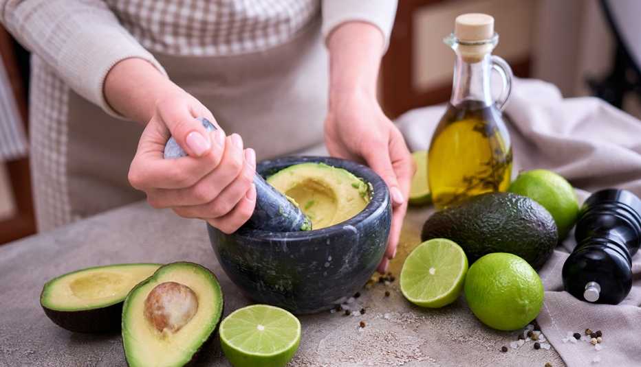 Woman using a mortar and pestle to prepare avocado puree on a kitchen counter with ingredients.